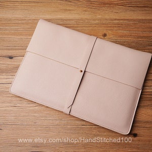 May include: A light pink leather laptop sleeve with a strap and a gold button closure. The sleeve is on a wooden surface.