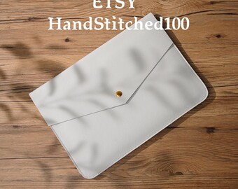 Könnte beinhalten: Eine weiße Ledertasche im Umschlag-Stil für Laptops mit einem goldenen Knopfverschluss. Die Tasche liegt auf einer Holzoberfläche. Der Text "ETSY HandStitched100" ist im Bild sichtbar.