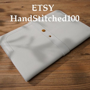 Puede incluir: Un portafolio de cuero gris claro con una superficie texturizada. El portafolio tiene un cierre de solapa con dos cierres a presi&oacute;n dorados. El texto "ETSY HandStitched100" est&aacute; impreso en el portafolio.