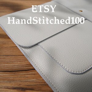 Puede incluir: Una funda de cuero gris claro estilo sobre con solapa y un broche dorado. La funda tiene una superficie texturizada y est&aacute; cosida con un hilo m&aacute;s claro. El texto "ETSY HandStitched100" est&aacute; impreso en la funda.