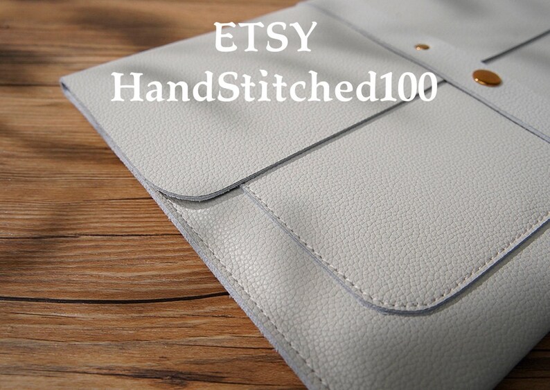 Puede incluir: Una funda de cuero gris claro estilo sobre con solapa y un broche dorado. La funda tiene una superficie texturizada y est&aacute; cosida con un hilo m&aacute;s claro. El texto "ETSY HandStitched100" est&aacute; impreso en la funda.