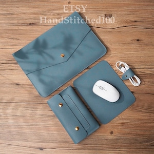 Puede incluir: Un conjunto de accesorios de cuero azul para portátil, que incluye una funda para portátil, una alfombrilla de ratón y una cartera. La alfombrilla de ratón tiene un ratón inalámbrico blanco encima. El conjunto está sobre una superficie de madera.