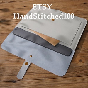 Puede incluir: Una funda para port&aacute;til de cuero gris claro con solapa y cierre de bot&oacute;n dorado. La funda tiene un bolsillo interior. El texto "ETSY HandStitched100" est&aacute; impreso en la parte superior.