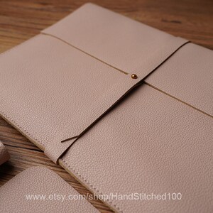 May include: A light pink leather laptop sleeve with a flap closure and a gold metal button. The sleeve is made of a textured leather and has a subtle sheen.