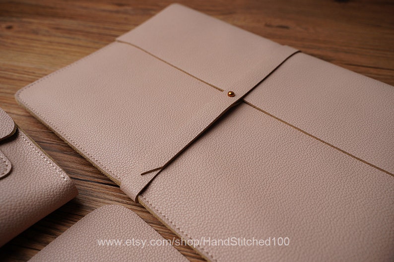 May include: A light pink leather laptop sleeve with a flap closure and a gold metal button. The sleeve is made of a textured leather and has a subtle sheen.
