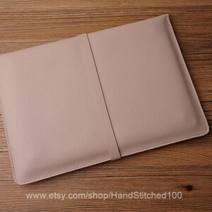 May include: A light pink leather laptop sleeve with a stitched seam down the center. The sleeve is lying on a wooden surface.
