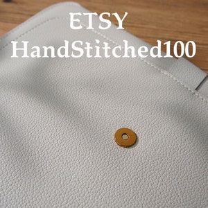 Puede incluir: Primer plano de un bolso de mano de cuero gris claro con una superficie texturizada. El bolso de mano tiene un cierre magn&eacute;tico dorado. El nombre de la tienda Etsy "HandStitched100" est&aacute; impreso en la parte superior.