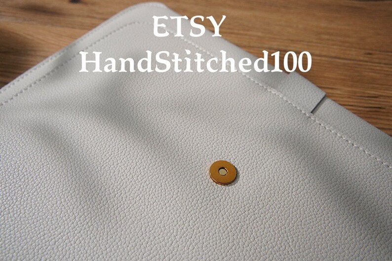 Puede incluir: Primer plano de un bolso de mano de cuero gris claro con una superficie texturizada. El bolso de mano tiene un cierre magn&eacute;tico dorado. El nombre de la tienda Etsy "HandStitched100" est&aacute; impreso en la parte superior.