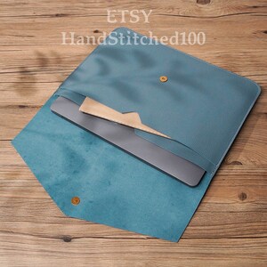 Op de afbeelding: Een tealblauwe leren laptophuls met een klep sluiting. De hoes is open en een laptop zit er in. De tekst "HandStitched100" is zichtbaar op de hoes.