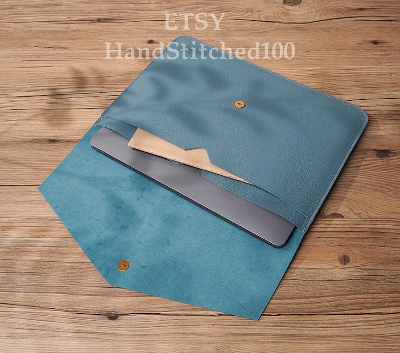 Op de afbeelding: Een tealblauwe leren laptophuls met een klep sluiting. De hoes is open en een laptop zit er in. De tekst "HandStitched100" is zichtbaar op de hoes.