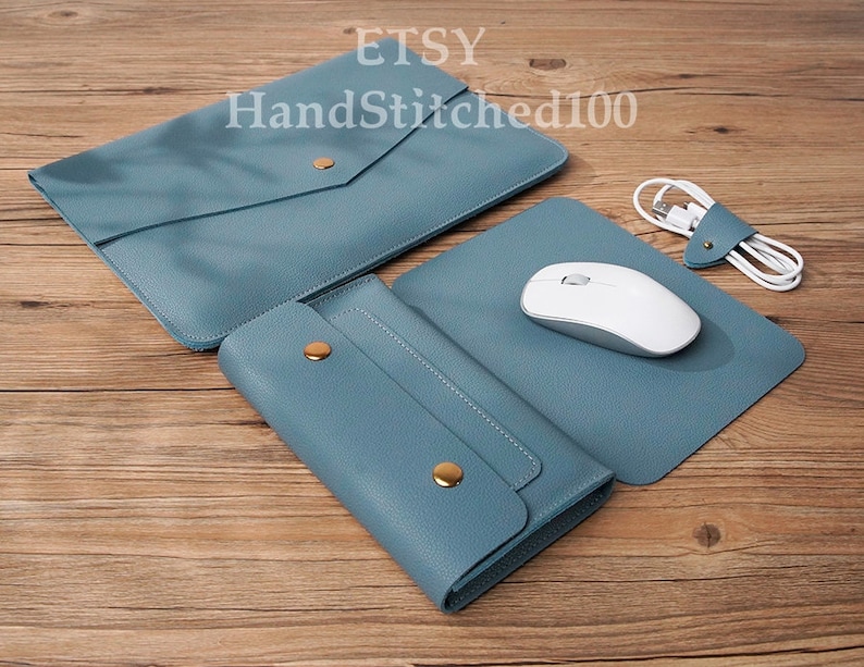 Op de afbeelding: Een set van drie blauwe leren accessoires: een laptophuls, een muismat en een portemonnee. De laptophuls heeft een klep met een gouden knoopsluiting. De muismat heeft een witte draadloze muis erop. De portemonnee heeft een gouden knoopsluiting en stikdetails. De tekst "Etsy HandStitched100" is zichtbaar op de laptophuls.