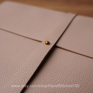 May include: A light pink leather folder with a gold metal closure. The folder has a textured surface and is folded in half.