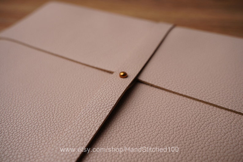 May include: A light pink leather folder with a gold metal closure. The folder has a textured surface and is folded in half.