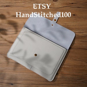 Puede incluir: Una funda para port&aacute;til de cuero gris claro con solapa y cierre de bot&oacute;n a presi&oacute;n. El interior es de un tono gris m&aacute;s claro. El nombre de la tienda Etsy "HandStitched100" est&aacute; impreso en la parte superior. La funda est&aacute; abierta.