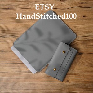 May include: Two gray leather pouches, one larger than the other, on a wooden surface. The larger pouch has a flap and the smaller pouch has a snap closure. Both pouches have gold-colored metal accents.