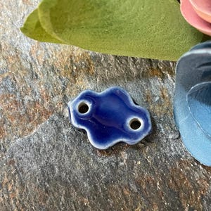 May include: A small, dark blue ceramic bead with two holes, shaped like a cloud. The bead has a glossy finish and a white edge. The bead is surrounded by a green leaf and a blue flower.