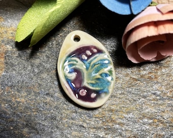May include: A ceramic pendant with a white background and a blue and purple design. The design features a stylized tree with a swirl pattern.