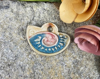 May include: A ceramic pendant with a unique design. The pendant features a light beige background with a blue and pink spiral design. The pendant has a hole at the top for hanging. The pendant is approximately 1.5 inches wide.