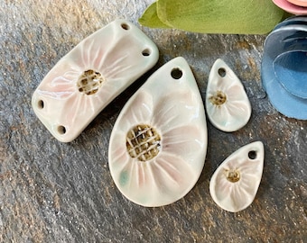 May include: A collection of ceramic flower-shaped pendants in various sizes. The pendants are white with pink petal details and a textured brown center. Each pendant has a hole for stringing. The background is a textured gray surface.