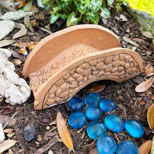 May include: A miniature terracotta bridge with a textured surface sits on a bed of brown mulch. Scattered around the bridge are ten small, round, blue glass stones.