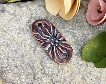 May include: A brown ceramic tile with a white floral design. The tile has two holes on the top and bottom.