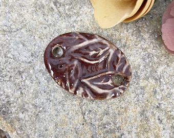 May include: A glossy, oval-shaped ceramic pendant in shades of burgundy and cream. The pendant features a leaf design and two holes for stringing. The pendant is approximately 2 inches long and 1 inch wide.