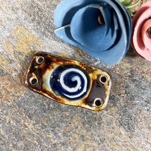May include: A rectangular ceramic pendant with a blue and white spiral design, framed by a brown border. The pendant has four holes for stringing. The background features blue and pink paper flowers on a gray stone surface.