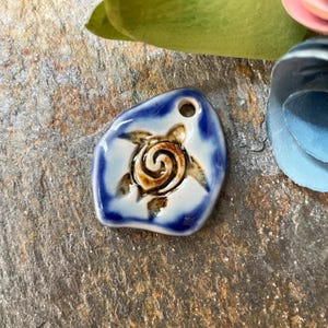 May include: A ceramic pendant with a turtle design. The pendant is a light blue irregular shape with a dark blue border. The turtle is brown with a spiral shell. A small hole is at the top for stringing.