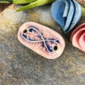 May include: A pink ceramic oval-shaped charm with a blue and white floral design. The charm has two holes on the side.