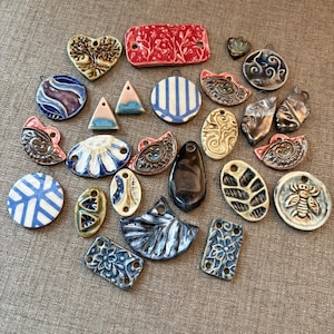 May include: A collection of various ceramic pendants and charms in diverse shapes, including hearts, circles, rectangles, and triangles. The pieces feature assorted glazes in blue, white, red, brown, and black, with embossed floral, bee, and swirl patterns.