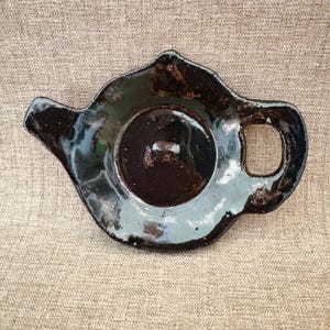 May include: A handcrafted, ceramic teapot-shaped tea bag holder. The holder is glazed in a dark, glossy brown with a light blue interior and a small handle. The edges have a speckled, rustic appearance. A unique addition to any tea set.