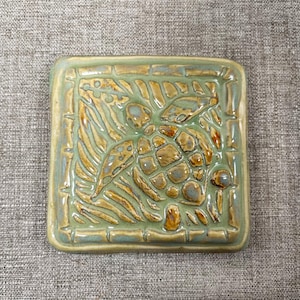 May include: Square ceramic tile with a raised turtle design. The tile features a green and brown color palette with a bamboo border. The turtle is surrounded by stylized leaves and other natural elements.