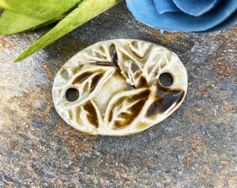 May include: A ceramic oval pendant with a brown leaf design. The pendant has two holes for stringing.