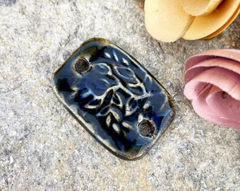 May include: A dark blue ceramic tile with a white floral design and two holes. The tile has a glossy finish.