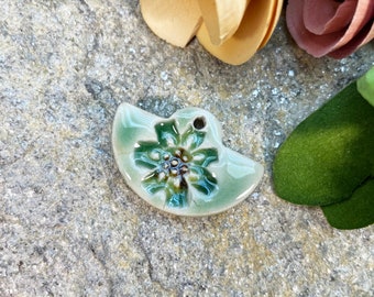 May include: A ceramic pendant in the shape of a half-circle with a green and brown floral design.