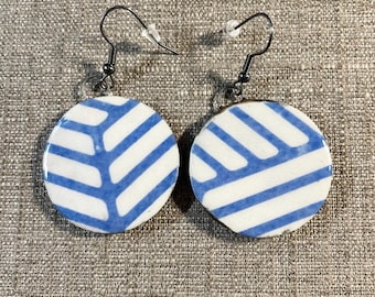 May include: A pair of round earrings with a white base and blue leaf-like patterns. The earrings have silver-colored hooks. The design is symmetrical, with the blue lines creating a visual texture. The earrings are displayed on a textured, neutral-colored surface.