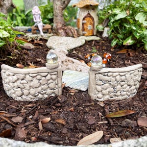 May include: A miniature stone wall with a rounded top and a small glass orb on each post. The wall is made of small, irregular stones and is painted a light brown color. The wall is set in a miniature garden with brown mulch and small green plants.