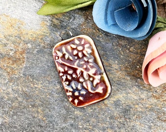 May include: A brown ceramic pendant with a white floral design. The pendant has a silver loop for hanging.