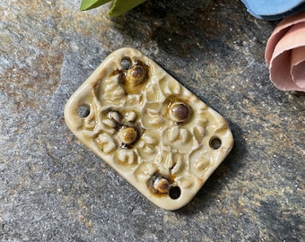 May include: A rectangular ceramic tile with a raised floral design. The tile is beige with brown accents and has two holes on the sides.