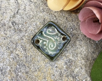 May include: A green ceramic tile with a white swirl design and two holes. The tile is in the shape of a diamond.