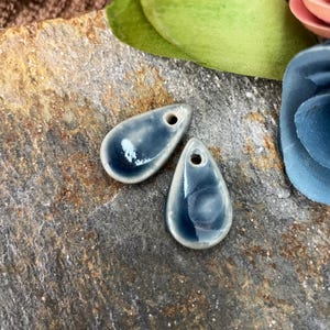 May include: Two teardrop-shaped ceramic pendants with a hole at the top for stringing. The pendants are a glossy, dark blue with a lighter blue swirl pattern. The edges are a light cream color. The pendants are displayed on a textured, gray stone surface.
