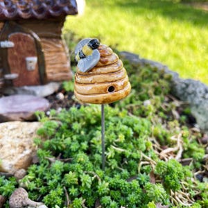 May include: A small ceramic beehive with a bee on top, painted in shades of brown, yellow, and black. The beehive is on a metal stake, which is inserted into the ground.