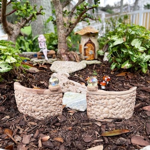 May include: A miniature fairy garden scene with a stone wall, a small house, and a path leading to it. The wall is made of small stones and has a rounded top. There are two small glass orbs on top of the wall, and a small fairy figurine is sitting on a bench in the background.