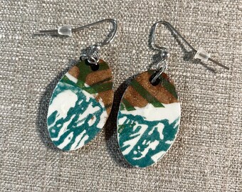 May include: A pair of oval-shaped earrings with a unique design. The earrings feature a combination of brown, green, white, and teal colors. The top portion has brown and green stripes, while the bottom has a teal and white pattern. The earrings have silver-colored hooks.