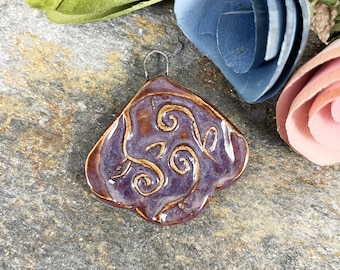 May include: A purple ceramic pendant with a swirl design in brown glaze. The pendant has a silver loop for hanging.