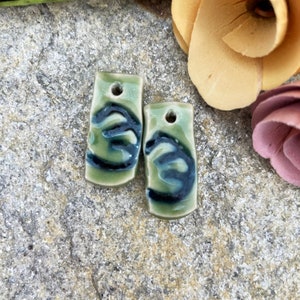May include: Two green ceramic rectangular tags with black swirl designs. Each tag has a hole at the top.