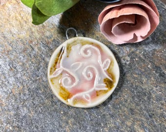 May include: A white ceramic pendant with a pink and brown glaze. The pendant features a swirling design with leaves and a loop for hanging.