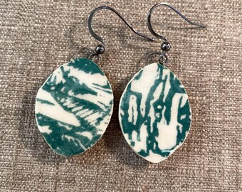 May include: A pair of handmade earrings with a unique design. Each earring features a white, oval-shaped ceramic pendant with a teal abstract pattern. The earrings have silver-colored ear wires and a small bead detail.