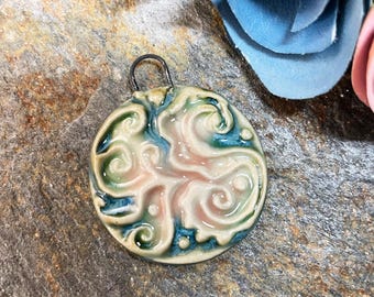 May include: A round ceramic pendant with a swirling design in shades of pink, green, and blue. The pendant has a silver loop for hanging.