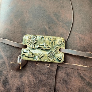 May include: A brown leather journal with a decorative clasp featuring sea life designs. The clasp is rectangular, with embossed images of fish, seashells, and a starfish. The journal has a leather strap closure.